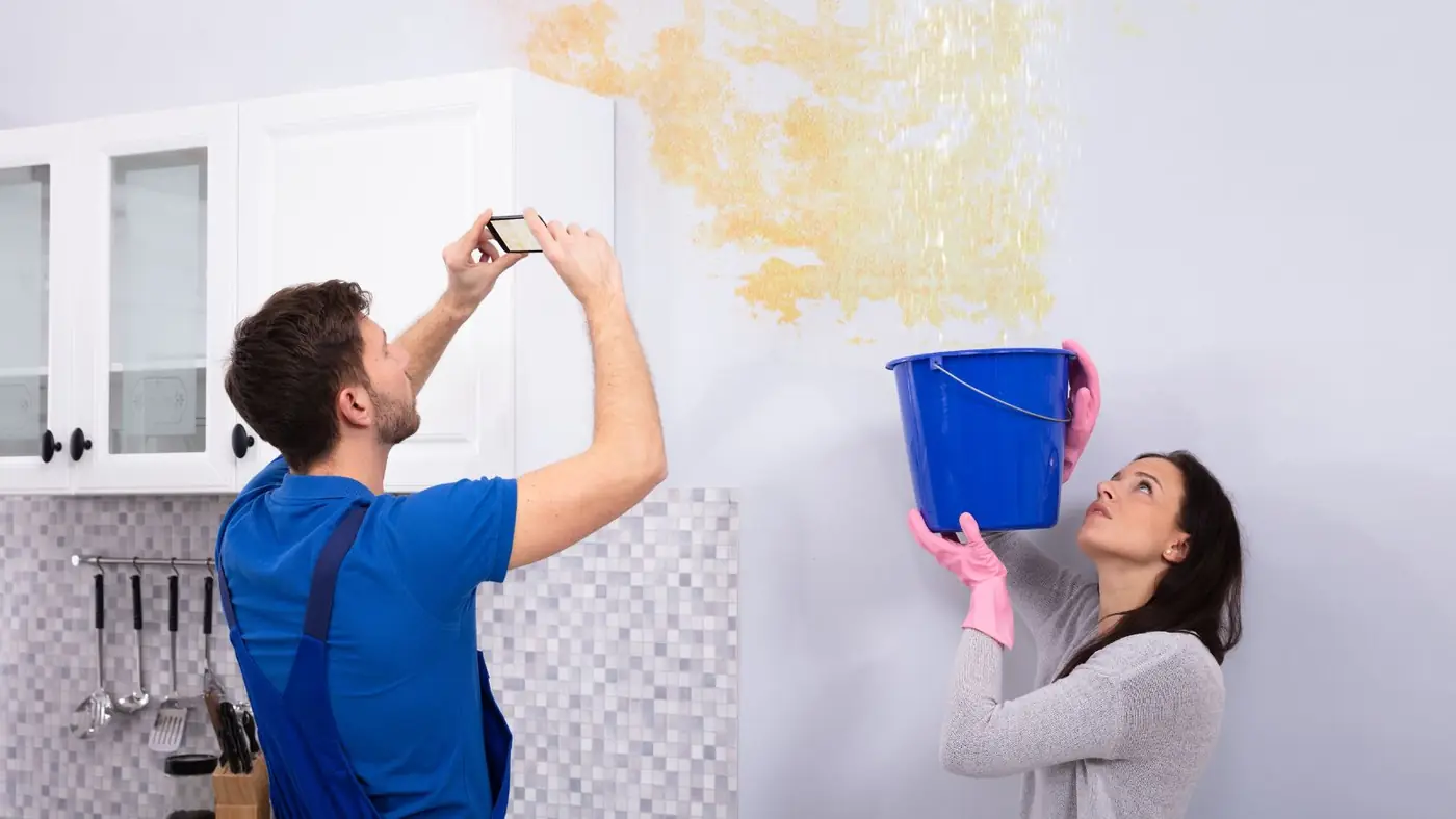 Man photographing a water-stained wall and ceiling with his phone while a woman holds a bucket below