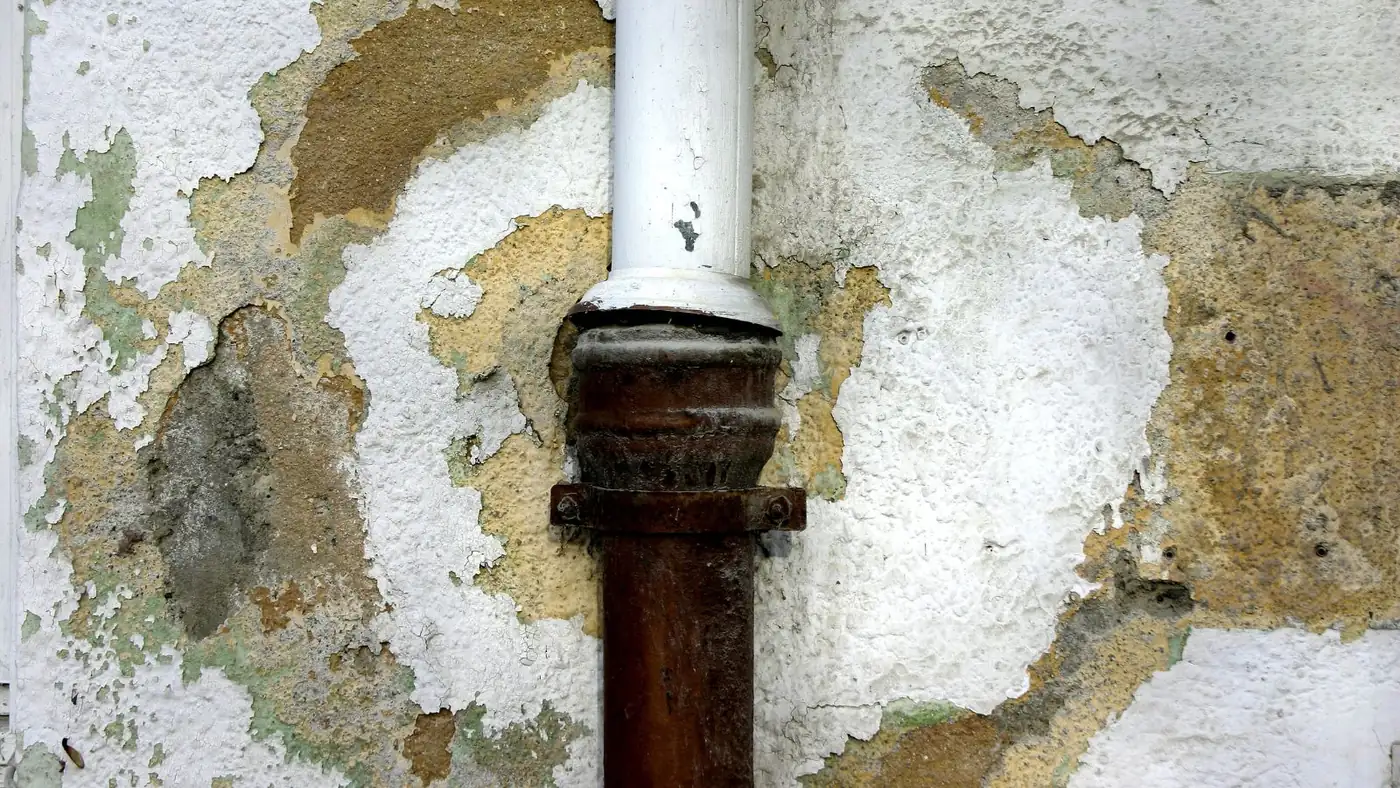 Water-damaged wall with peeling paint and stained plaster around a rusted vertical pipe